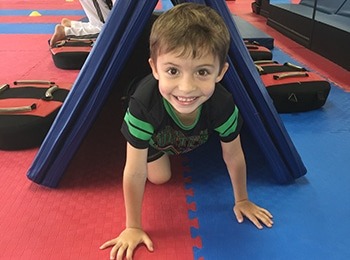 A smiling young boy crawling through a blue mat tunnel during an obstacle course at a fun and safe March break camp Burlington.