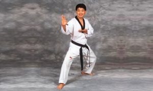 A male martial arts instructor in a white Taekwondo uniform with a black belt performing a defensive hand strike stance.