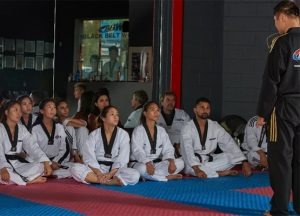 A group of diverse Taekwondo students in white uniforms and black belts sitting cross-legged on a training mat, listening to an instructor in a black uniform.