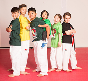 A diverse group of young children in colorful t-shirts and Taekwondo pants posing playfully together on a red training mat.