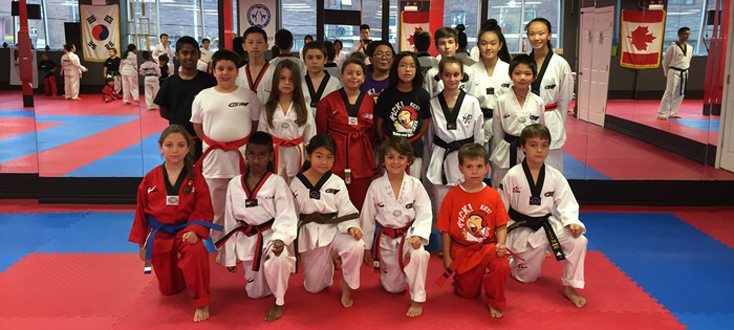A large group of children in white uniforms and various colored belts practicing punching techniques on red and blue mats in a spacious dojang.