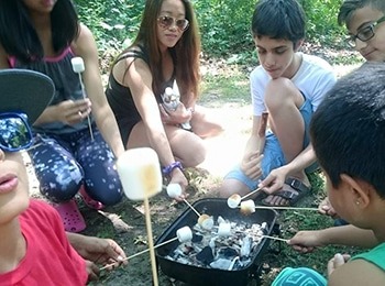 A group of young campers and an instructor sitting together outdoors roasting marshmallows over a small charcoal grill during a summer day camp session.