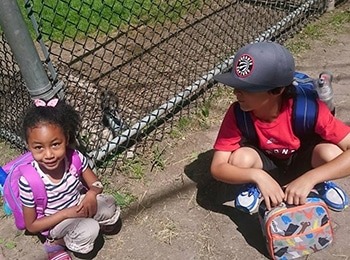A young girl and boy with backpacks and lunch bags crouching together by a fence outdoors during a local martial arts summer camp session.